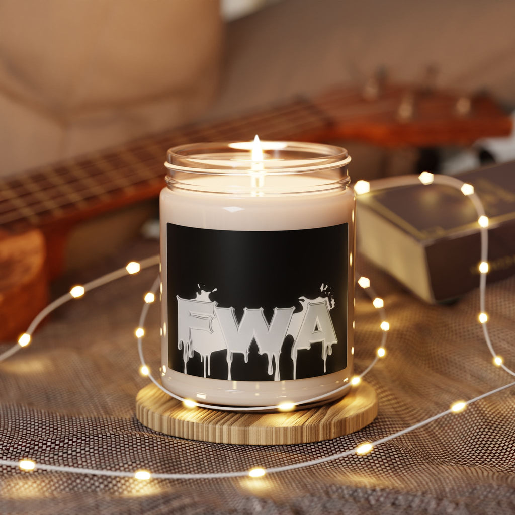 9oz Scented Soy Candle in a glass jar with a black label featuring a white 'FWA' drip logo, burning on a wooden coaster with fairy lights and a ukulele in the background.